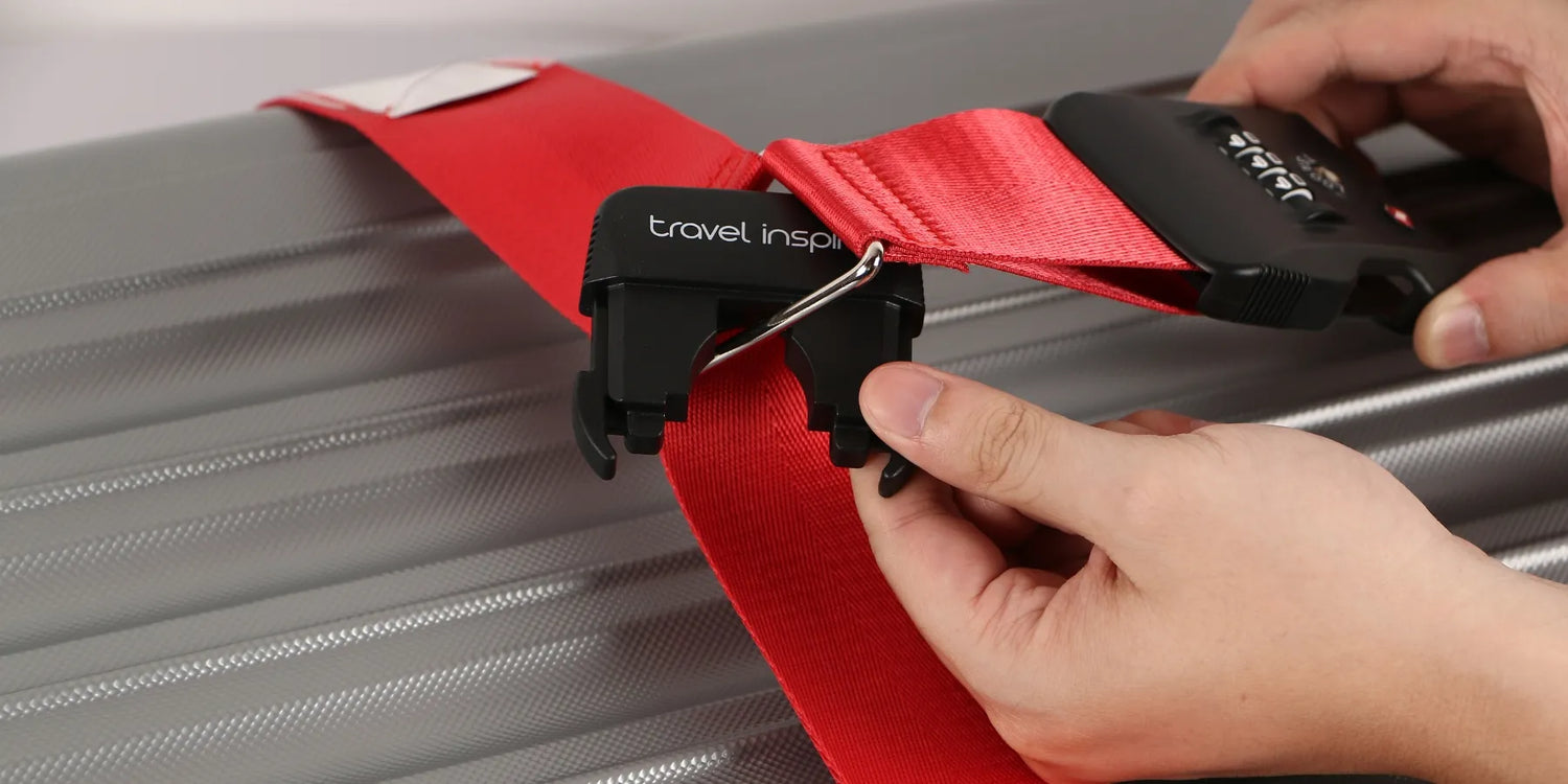 A close-up of hands securing a red Travel Inspira luggage strap with a combination lock onto a gray suitcase. The strap is being fastened using a black buckle with the brand name "travel inspira" clearly visible. This luggage strap is a key travel accessory for securing suitcases.