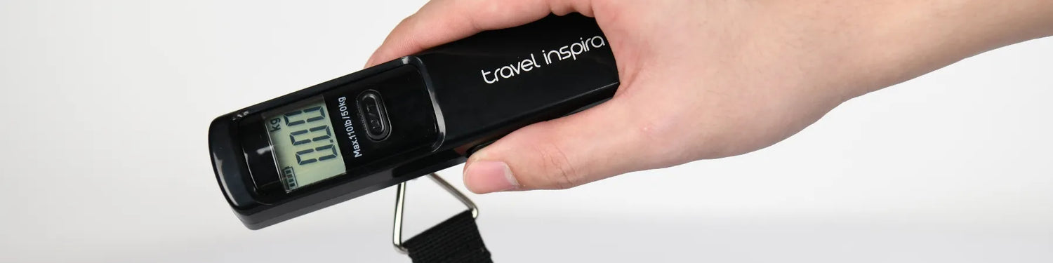 A hand holding a black Travel Inspira digital luggage scale. The scale displays "00.00 kg" on its screen. It is a portable luggage weight scale, perfect for travel accessories.