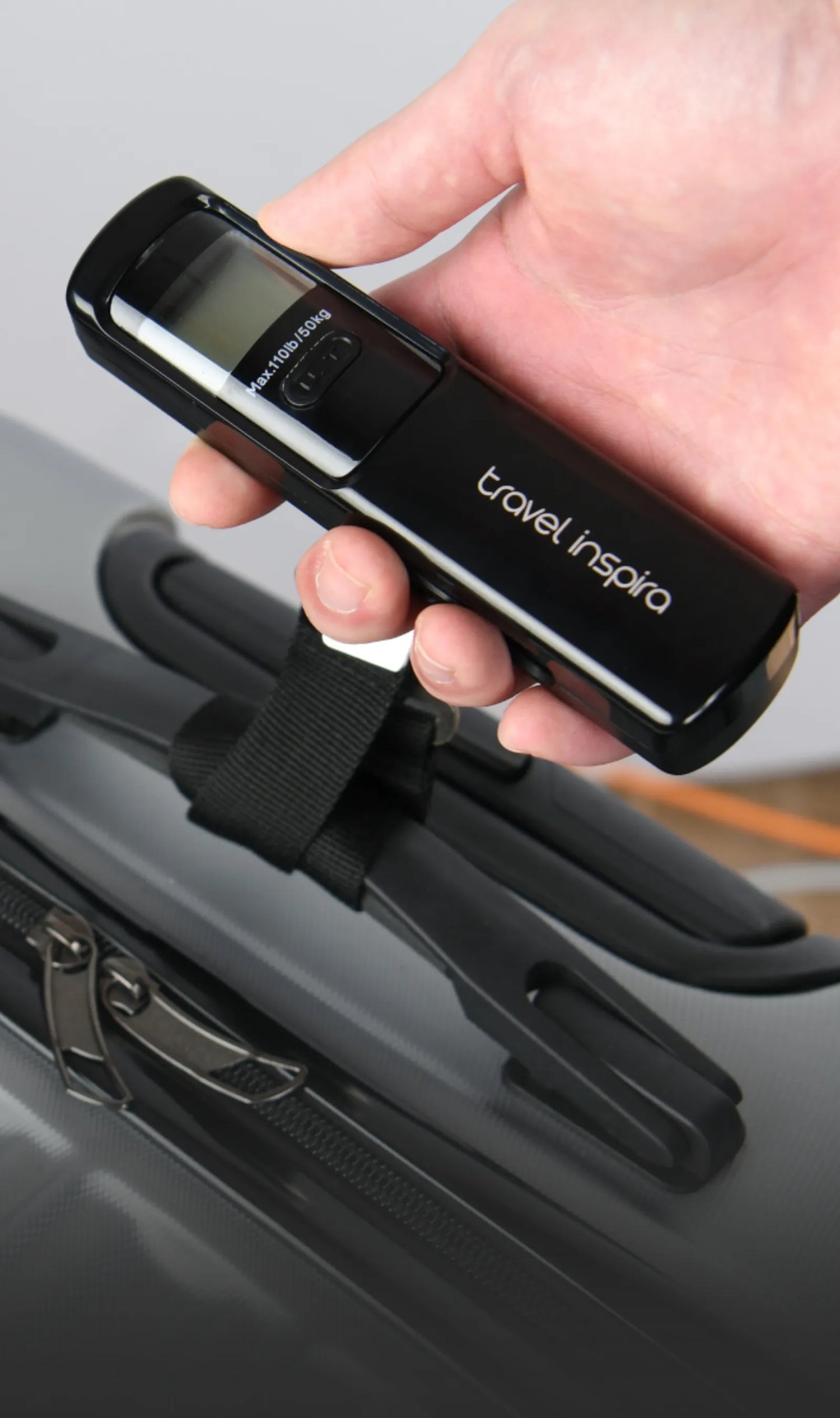 A hand holding a black Travel Inspira digital luggage scale with a display screen. The scale has a maximum weight capacity of 110lb/50kg. It is positioned above a suitcase, highlighting its use as a travel accessory. Keywords: Travel Inspira, Digital Luggage Scale, Luggage Weight Scale, Travel Accessories