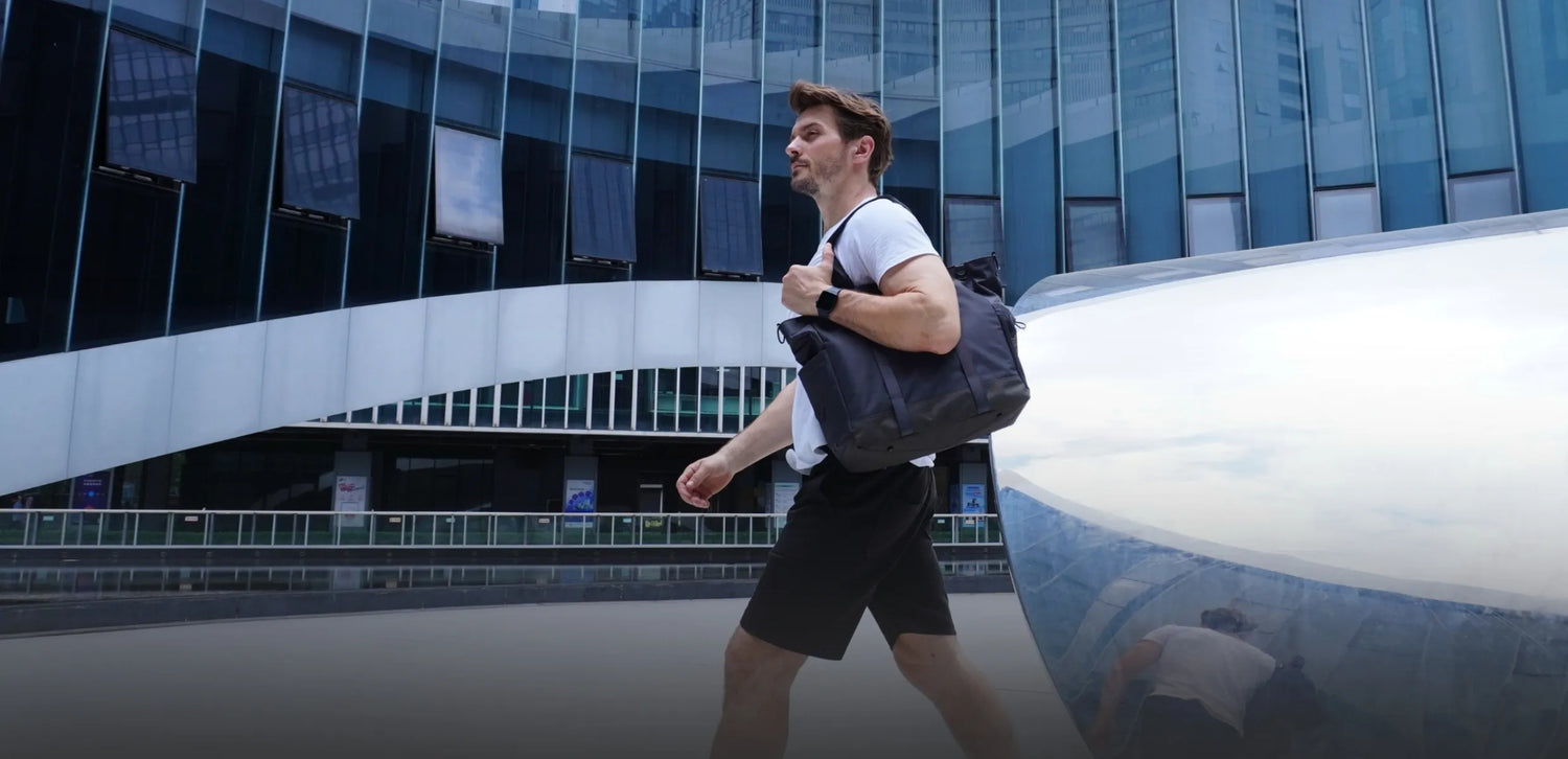 A man wearing a white t-shirt and black shorts is walking outdoors, carrying a black travel backpack over his shoulder. The background features a modern building with large glass windows. This image is relevant to travel accessories, specifically highlighting the use of a backpack for travel. Keywords: Travel Backpack, Travel Accessories, Modern Building, Outdoor Travel