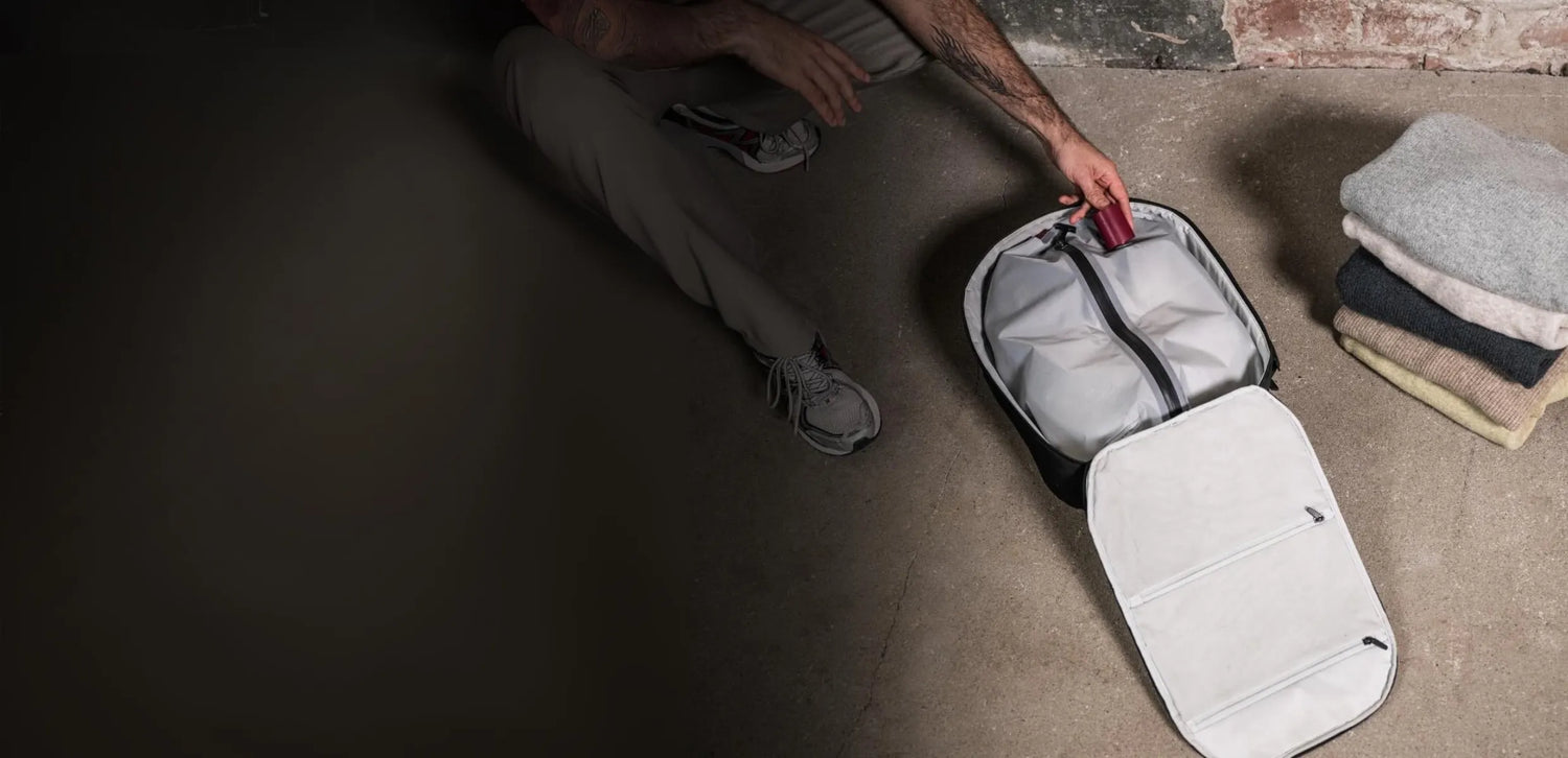 A person is packing clothes into a Travel Inspira luggage. The luggage is open, revealing its spacious interior. The person is placing a small red item, possibly a battery-free luggage scale, into the luggage. There are folded clothes stacked beside the luggage. This image showcases travel accessories, specifically focusing on the use of a luggage scale during packing.