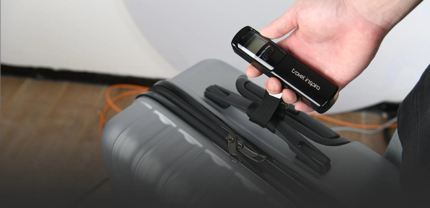 A hand holding a black Travel Inspira luggage scale over a gray suitcase. The scale is being used to measure the weight of the luggage. Keywords: Travel Inspira, Luggage Scale, Suitcase, Travel Accessories