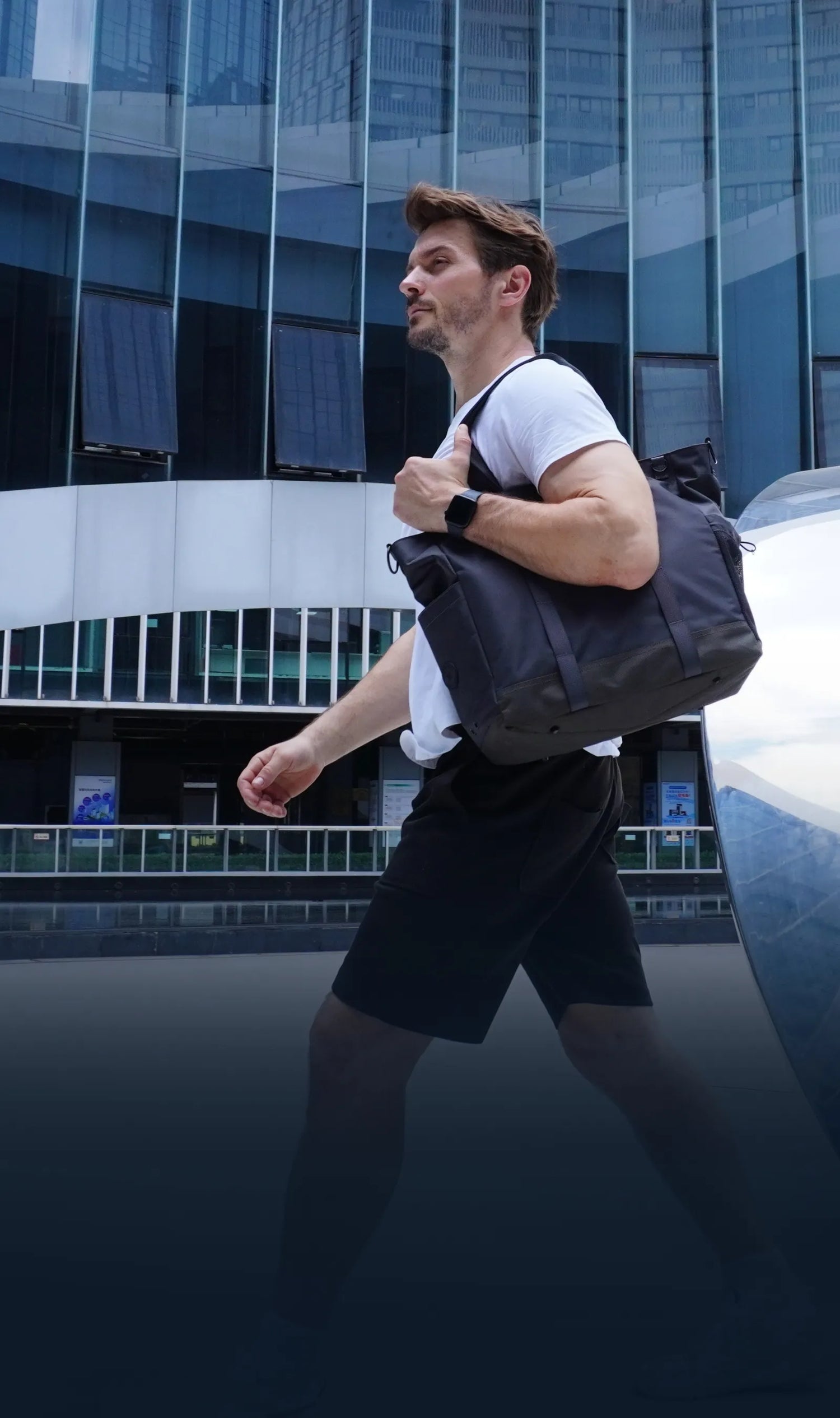 A man wearing a white t-shirt and black shorts is carrying a Travel Inspira travel bag over his shoulder. He is walking in front of a modern building. The bag is suitable for travel, and the brand Travel Inspira is known for travel accessories.