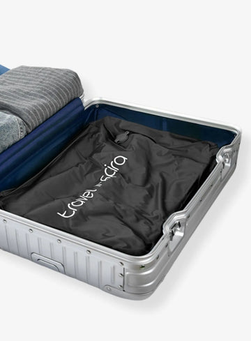 Open silver suitcase with blue interior. Inside, neatly folded clothes (gray striped shirt and denim) and a black Travel Inspira branded travel vacuum compression bag. Ideal for organizing luggage. Keywords: Travel Accessories, Travel Inspira, Vacuum Compression Bag
